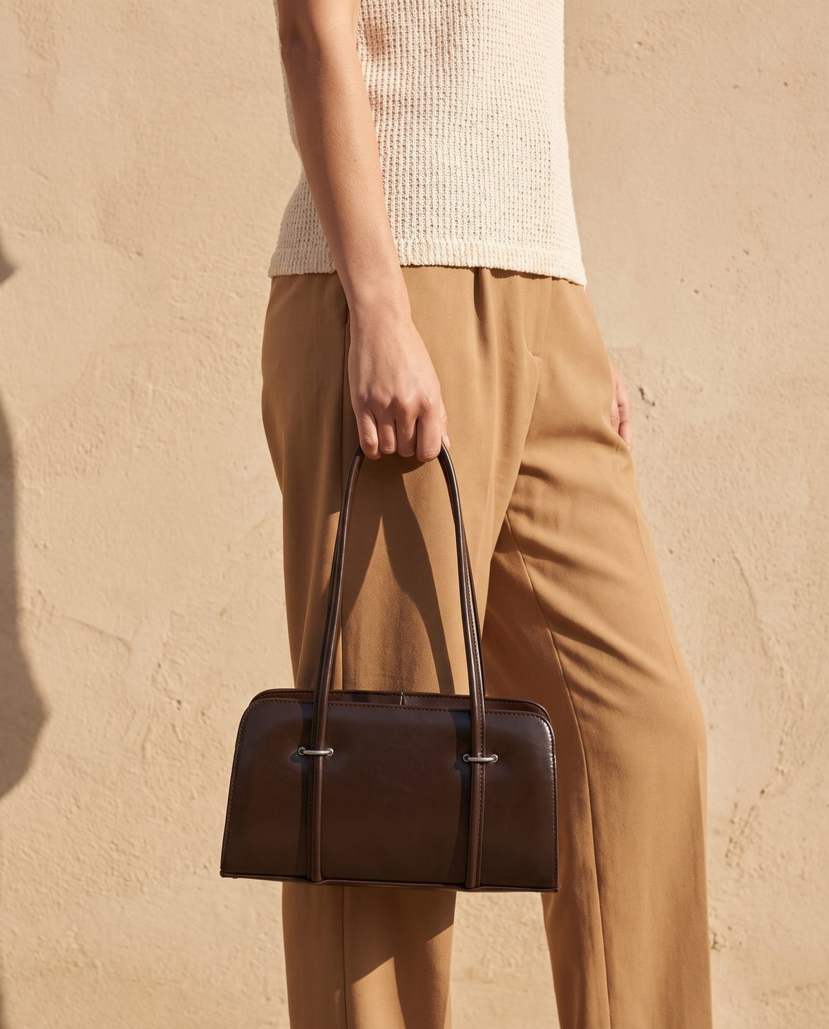 Model in a cream knit top and tan trousers holding the Badr espresso brown bag against a sand-coloured wall – Nouva