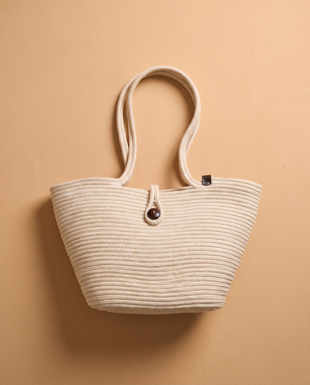 A cream-colored woven straw tote bag with dual shoulder straps and a central dark brown button closure, displayed against a solid warm beige background.