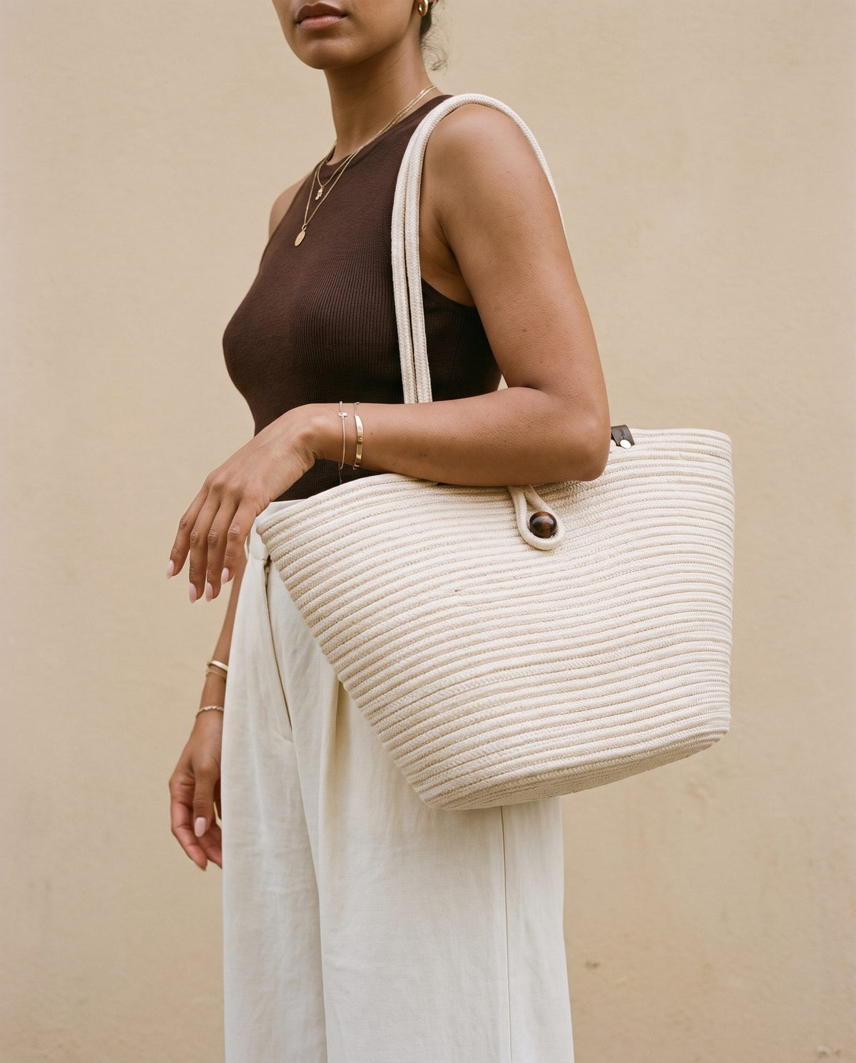 A woman wearing a dark brown sleeveless top and white wide-leg pants carries a large cream-colored woven straw tote bag with a white shoulder strap against a beige wall.