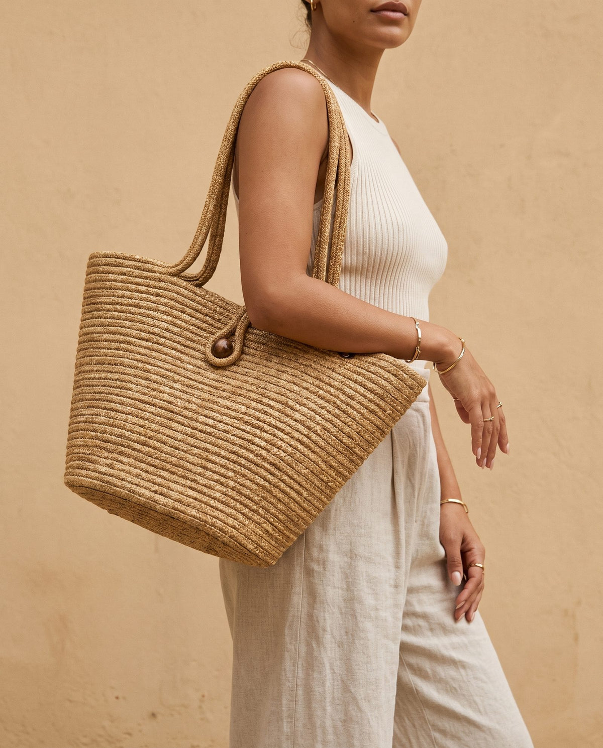 A woman wearing a white ribbed tank top and light linen pants carries a large woven straw tote bag with double shoulder straps against a warm beige stucco wall.