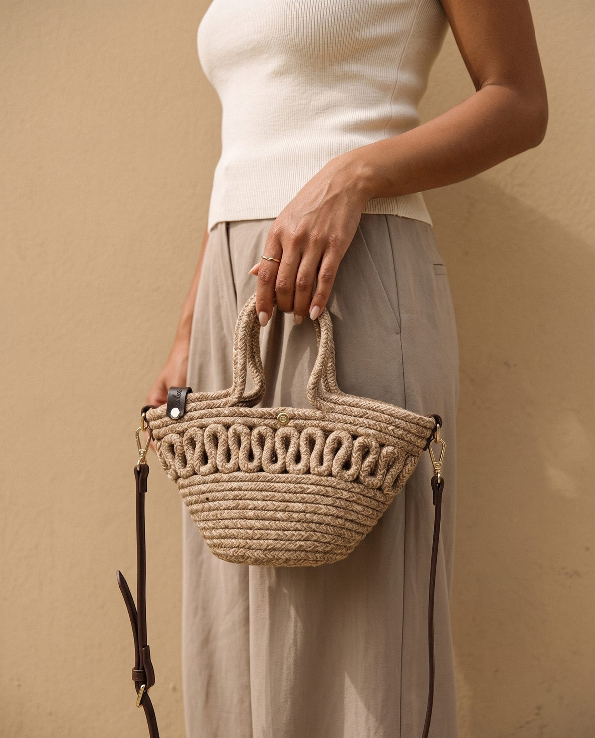 Model in a cream ribbed top and taupe trousers holding the Kawkab Mini Straw Crossbody Bag in taupe by its top handles against a warm sand wall – Nouva