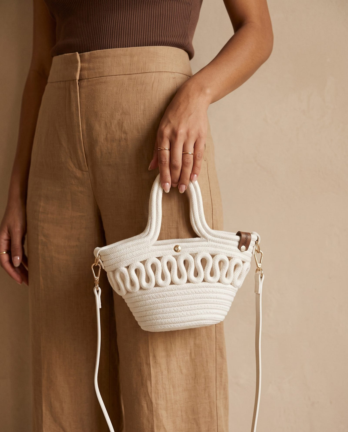 A person wearing tan trousers and a brown ribbed top holds a small structured woven handbag with white rope-like material, double top handles, gold-tone hardware, and a long matching strap against a warm beige wall.