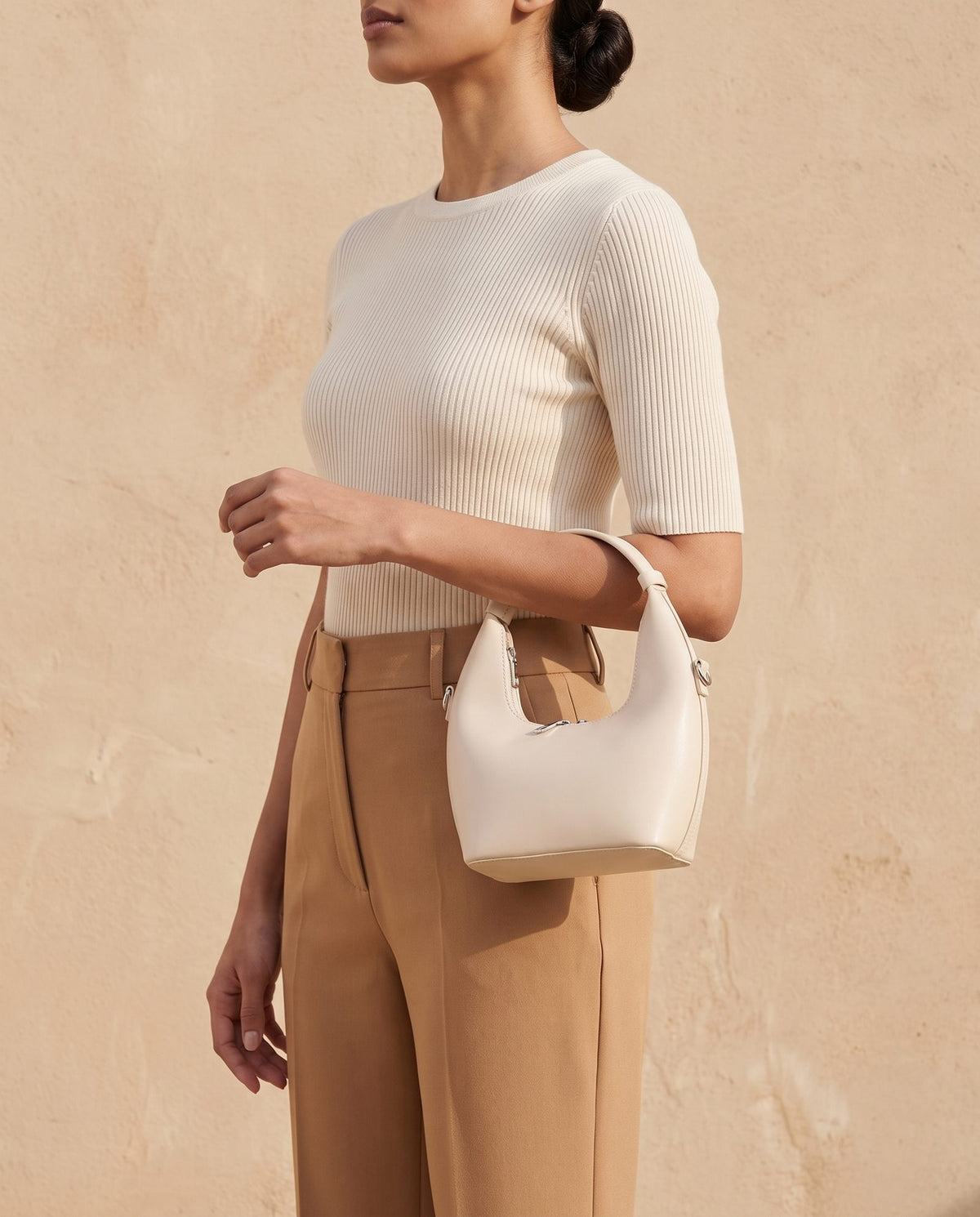 Model in a cream knit top and camel trousers holding the Mizar cream bucket bag by its short strap, compact 20 cm women's bag by Nouva