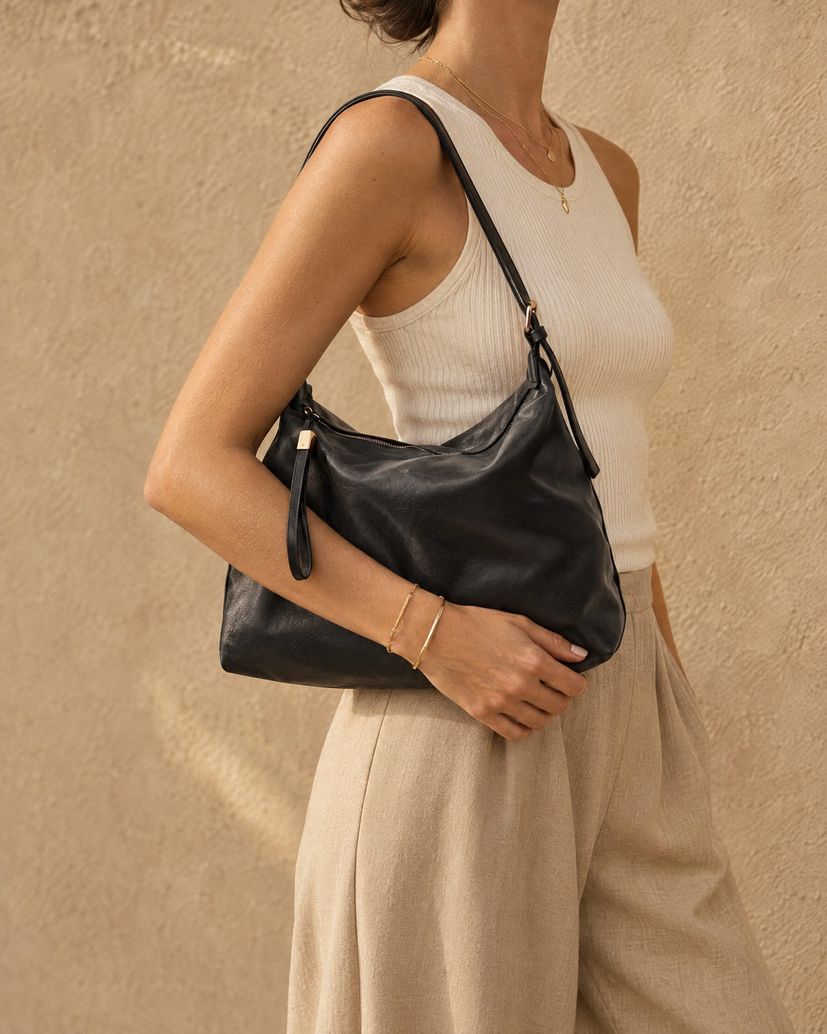 A woman wearing a cream ribbed tank top and beige wide-leg pants carries a black shoulder bag against a textured beige wall.