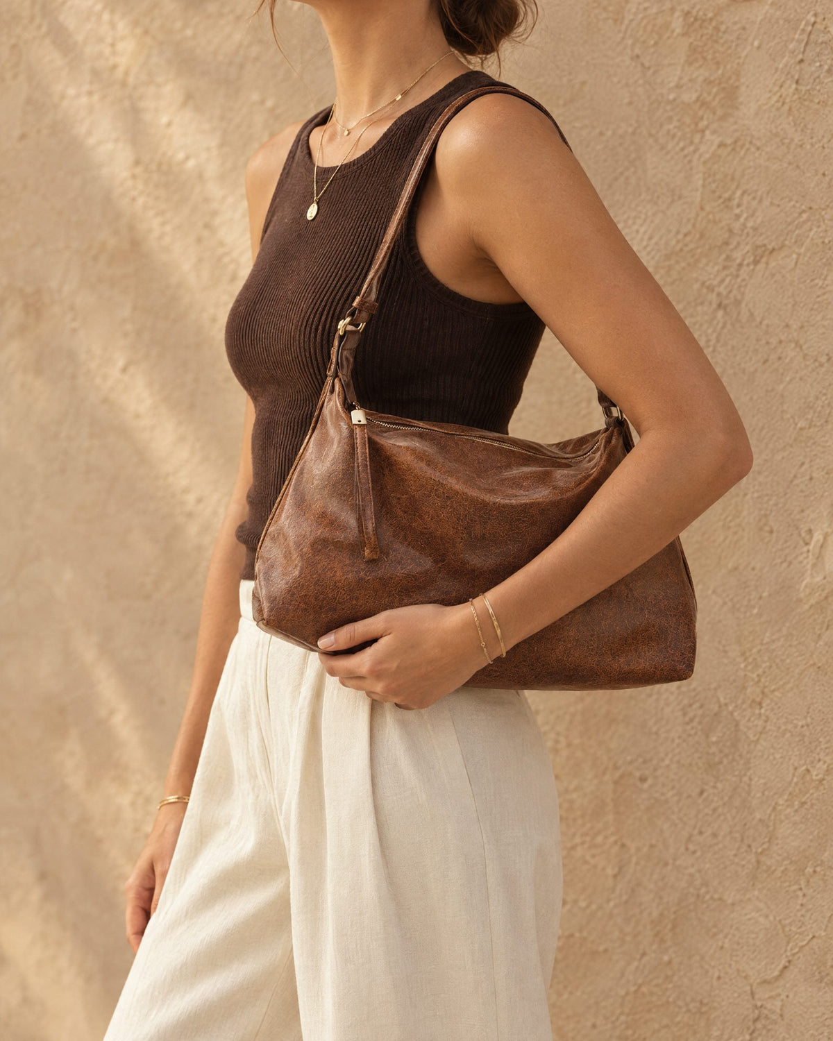 A woman wearing a dark brown ribbed tank top and white wide-leg pants carries a large distressed brown shoulder bag against a textured beige stucco wall.
