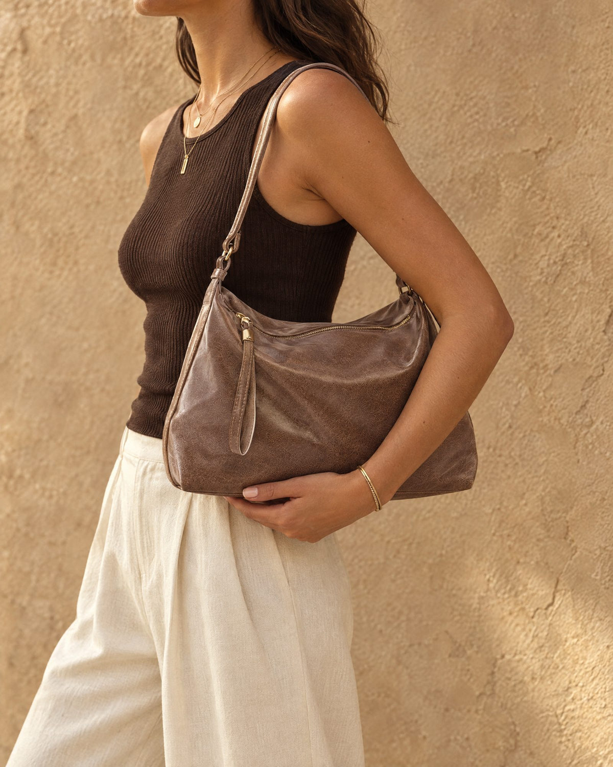 Model in a dark brown ribbed tank top and cream trousers carrying the Qamar Slouchy Shoulder Bag in taupe on her shoulder against a textured sand wall – Nouva